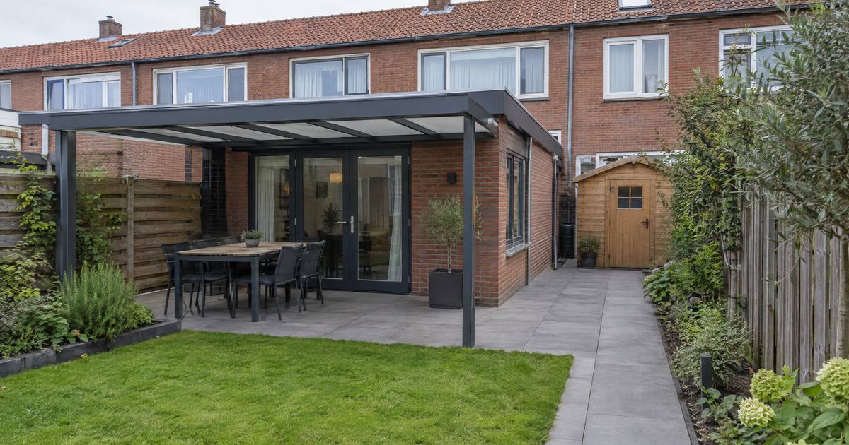 Een achtertuin achter een rijtjeshuis van rode baksteen, met een uitbouw tegen het huis, een houten schuur achterin en een overkapping boven het terras. Illustratie.